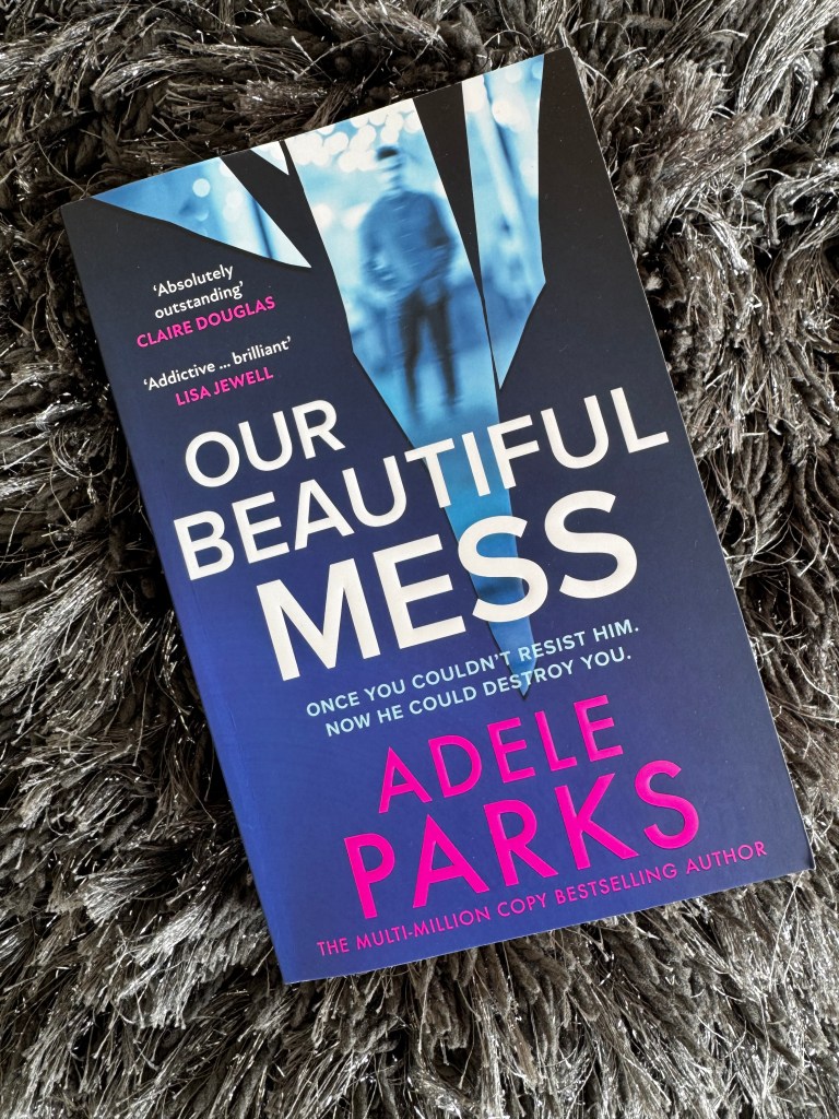 A close-up of the book 'Our Beautiful Mess' by Adele Parks, featuring a striking blue and black cover design with bold pink text, resting on a textured gray surface.